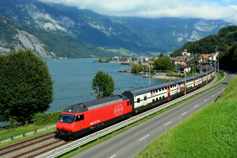 Red SBB Re 460 locomotive hauling an IC 2000 double-deck InterCity train along a Swiss lakeshore, the kind of long-distance trip behind the Swiss Travel Pass vs Half Fare Card decision