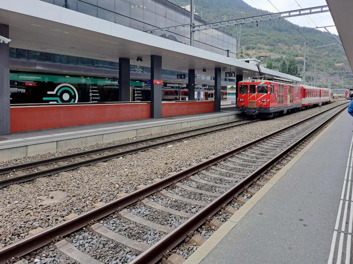 Railway platform at Visp station with a red train and Alpine slopes behind